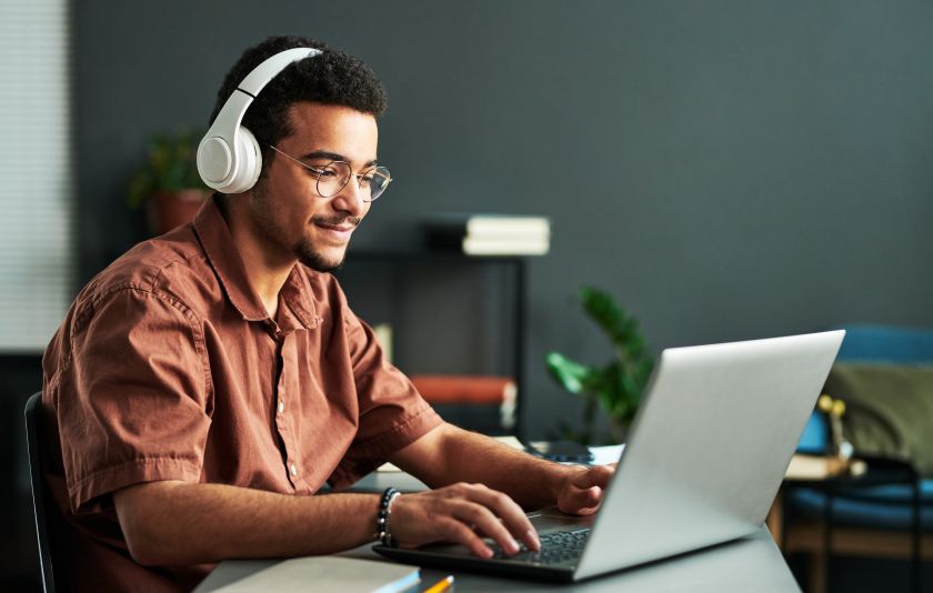 A young Black man wearing glasses and headphones and smiling while using his laptop. 