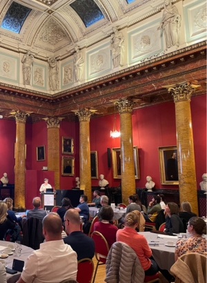 Image of the VCCP workshop held in Edinburgh. Shows a speaker at a lectern in  a red room with an audience sat around circular tables facing the speaker.